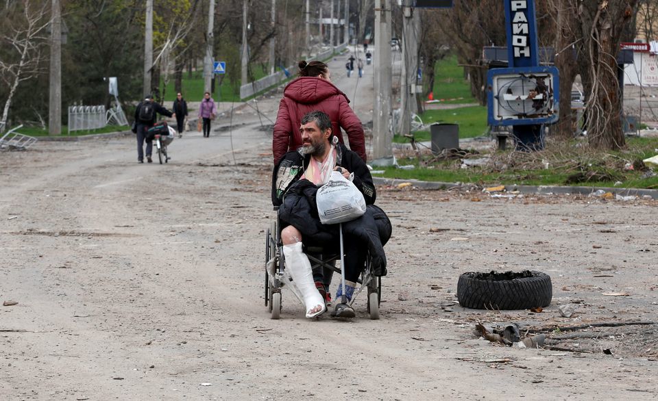 Một phụ nữ đẩy chiếc xe lăn chở một người đ&agrave;n &ocirc;ng bị thương tr&ecirc;n đường phố trong cuộc xung đột Ukraine-Nga ở th&agrave;nh phố cảng Mariupol, miền nam Ukraine ng&agrave;y 18/4/2022. Ảnh: Reuters